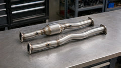 Catted and catless stainless-steel downpipes displayed side by side on an automotive workshop bench.